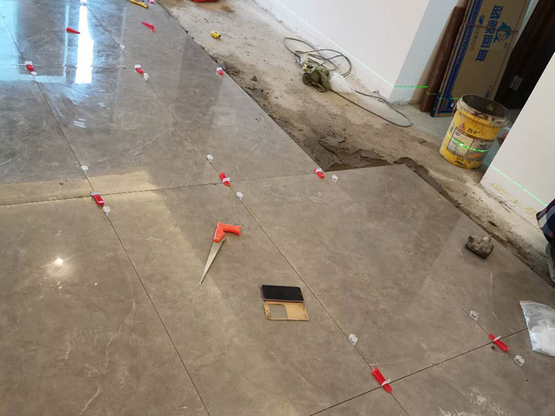 客廳地磚鋪貼：地磚鋪貼前，應(yīng)將地面清掃干凈，尤其是凝結(jié)后的混凝土，否則，地磚鋪貼后會(huì)出現(xiàn)翹邊、空鼓等現(xiàn)象，然后再灑水潤(rùn)濕地面。地面充分濕潤(rùn)，但不能有積水，然后將3:1混合好的沙子水泥鋪在地面上，進(jìn)行基層處理。在地磚背面均勻抹水泥砂漿作為粘合層，厚度不低于1cm，然后鋪在剛才試鋪的位置上，用橡皮錘輕敲地磚飾面直至密實(shí)平整達(dá)到要求。地磚鋪完后24h進(jìn)行清理勾縫，勾縫前應(yīng)先將地磚縫隙內(nèi)雜質(zhì)擦凈，用專用填縫劑勾縫，完工后（一般宜在24h之后）再用棉紗等物對(duì)地磚表面進(jìn)行清理。